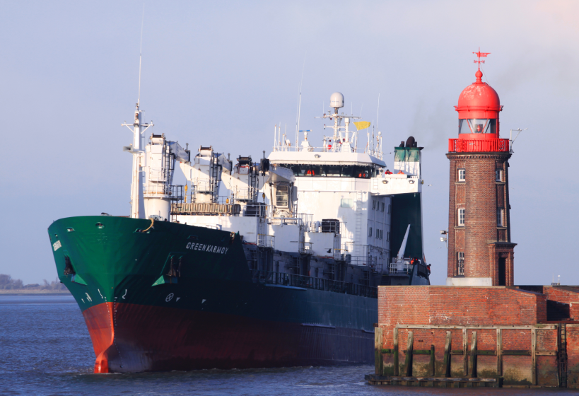 Grünes Frachtschiff „Green Karmoy“ neben einem roten Leuchtturm auf dem Wasser.