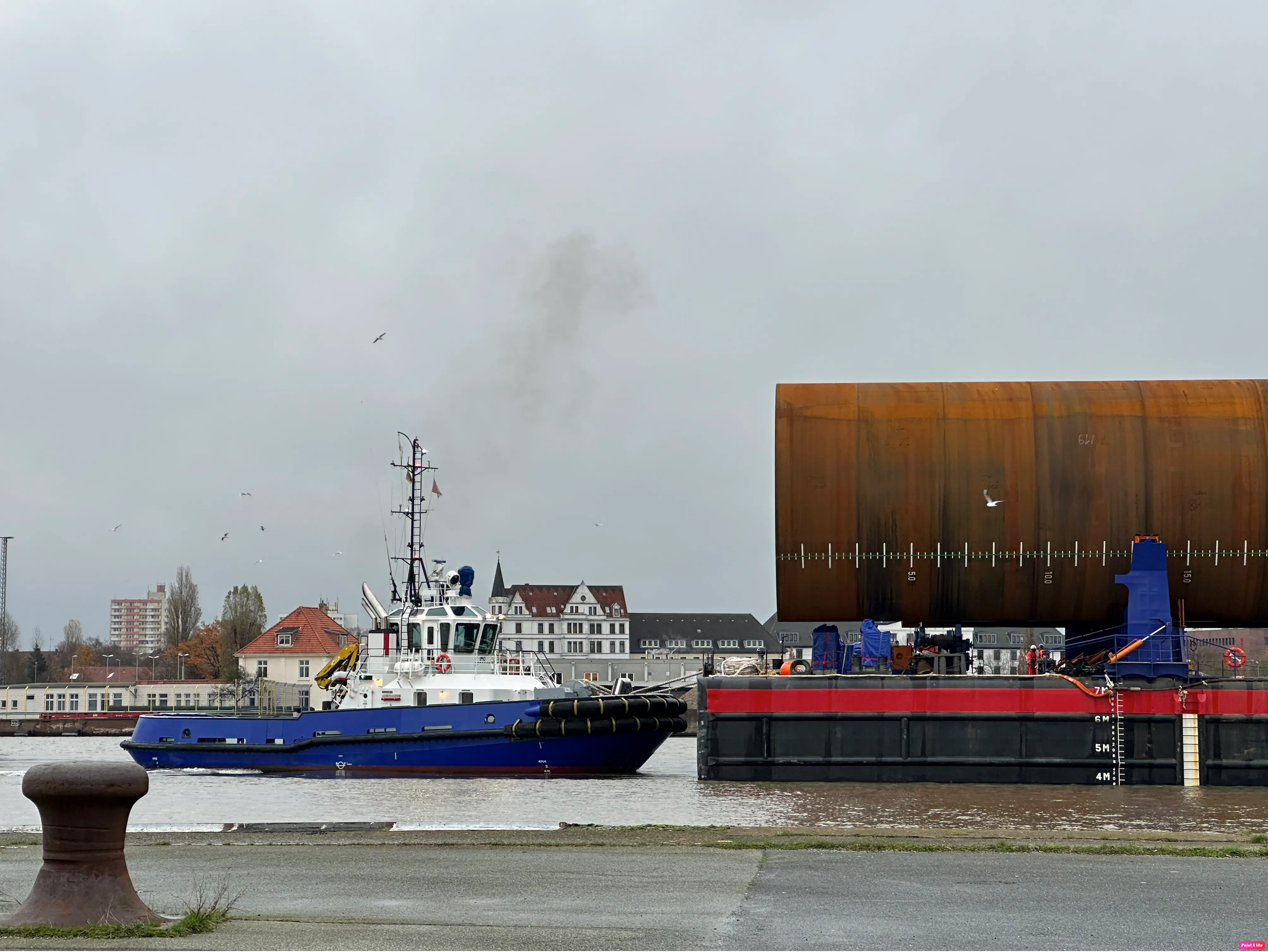 Schlepper zieht eine große Last mit rostbrauner Struktur durch einen Hafenbereich.
