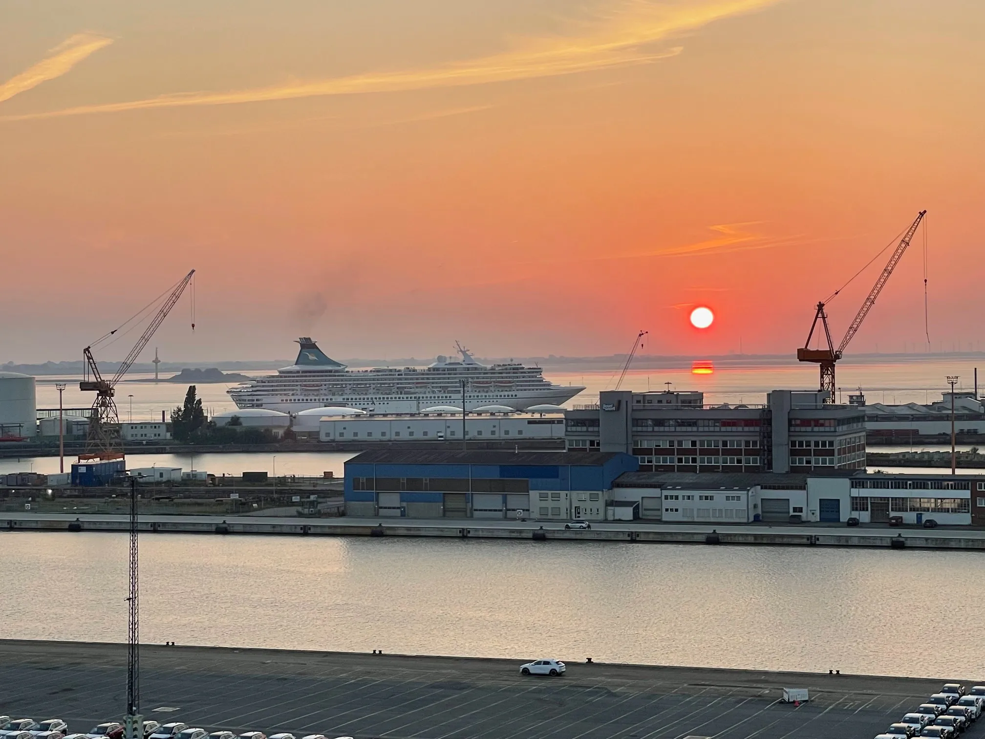 Sonnenuntergang im Hafen mit einem Kreuzfahrtschiff und Kränen vor Wasserfläche.